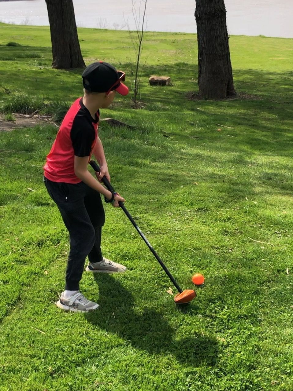 Child playing golf on a grassy field, wearing a red shirt and black cap, aiming at an orange ball.