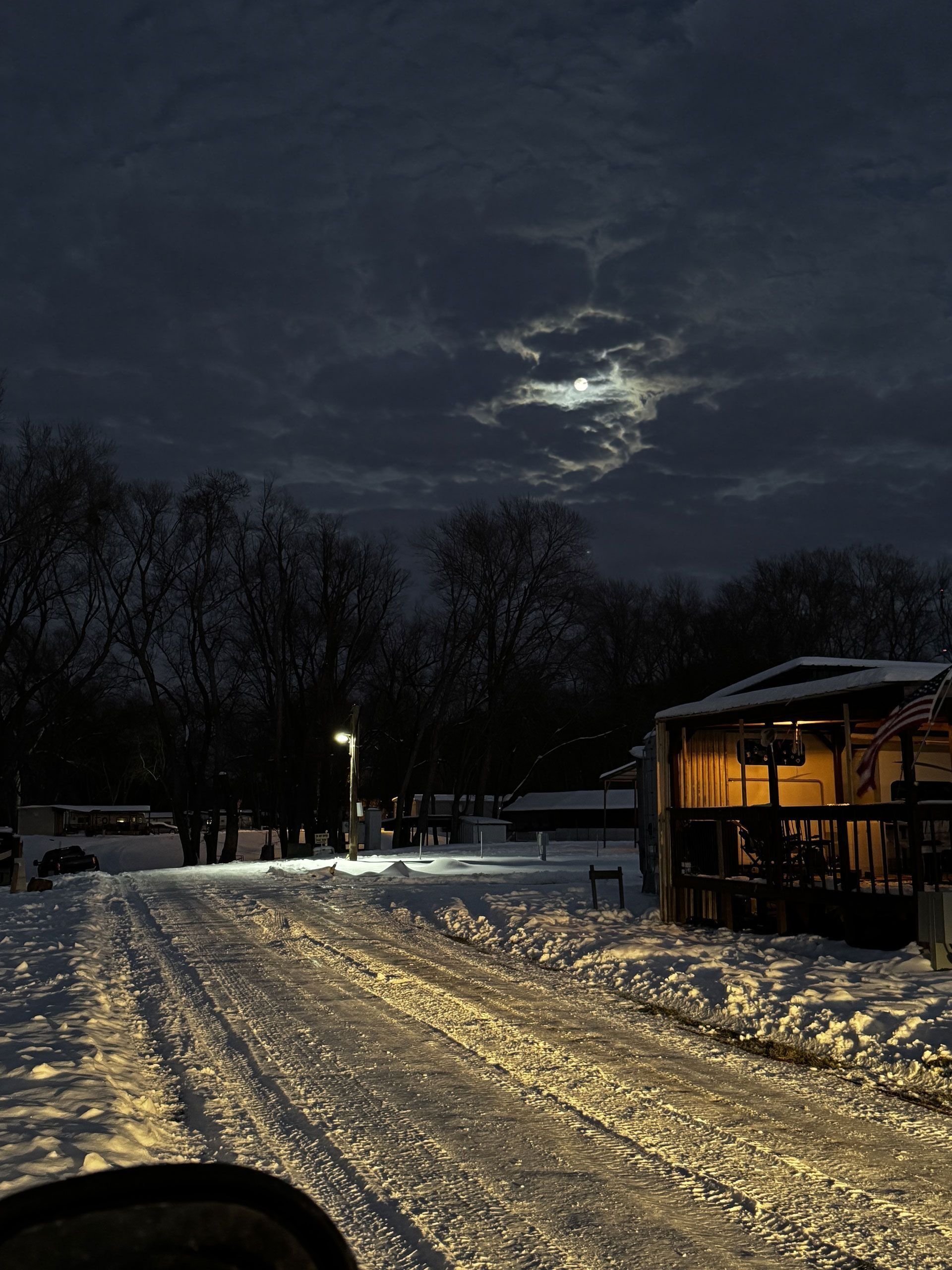 Snowy road at night with streetlights, bare trees, and a lit cabin under a cloudy moonlit sky