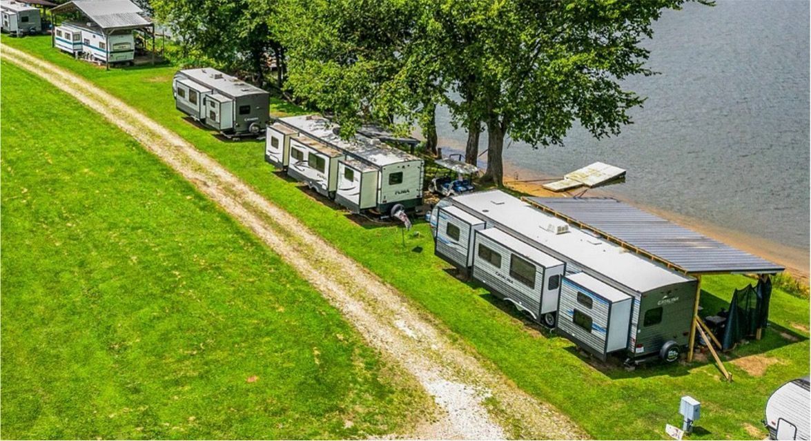 RVs parked on a grassy lakeside campground beside calm water, with a dirt path and dock nearby