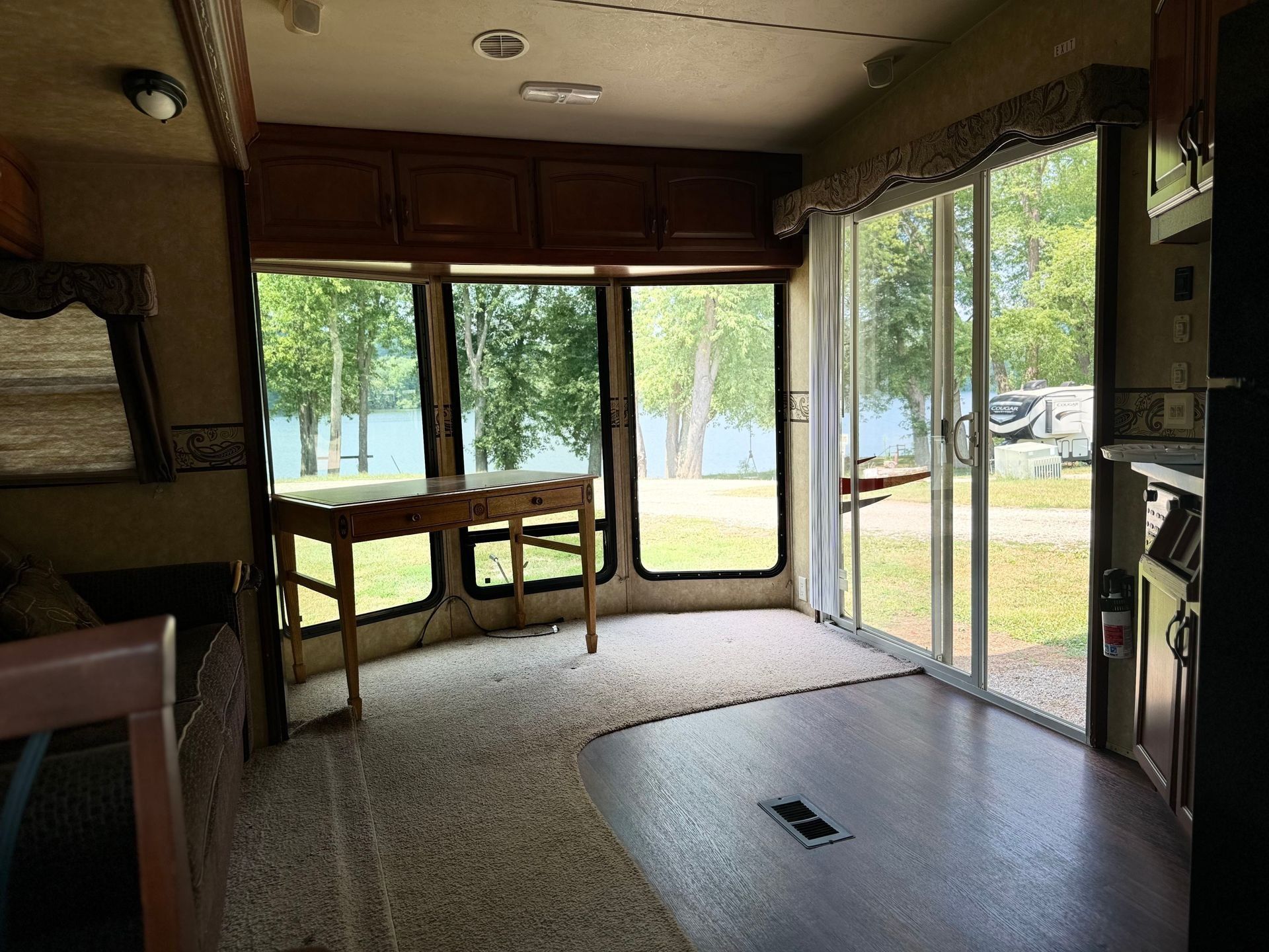 Empty sunroom with large windows, wood table, carpeted floor, and a view of trees and parked cars.