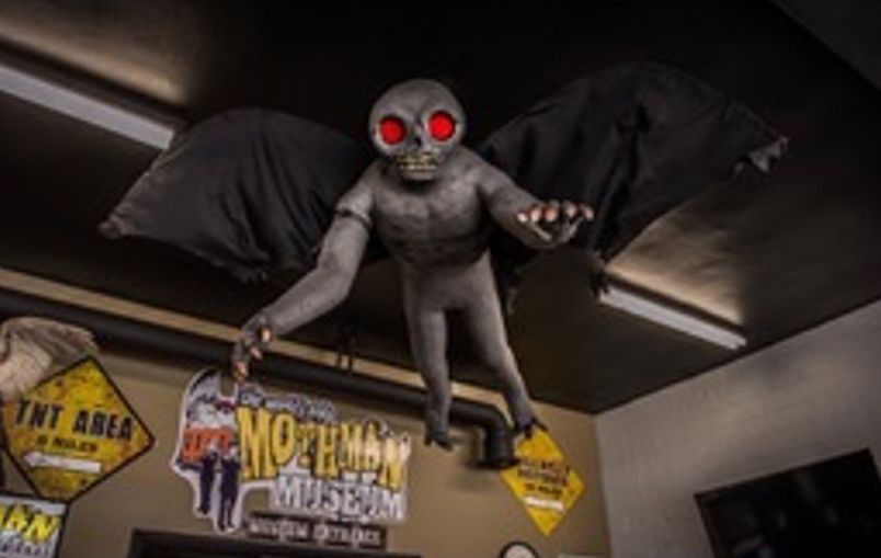 Grey creature with glowing red eyes perched on a ceiling, over a wall with vintage signs.