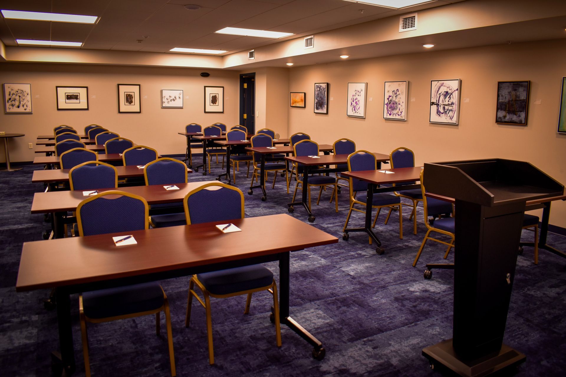 Empty conference room with tables, chairs, and a podium. Blue carpet, tan walls with artwork.