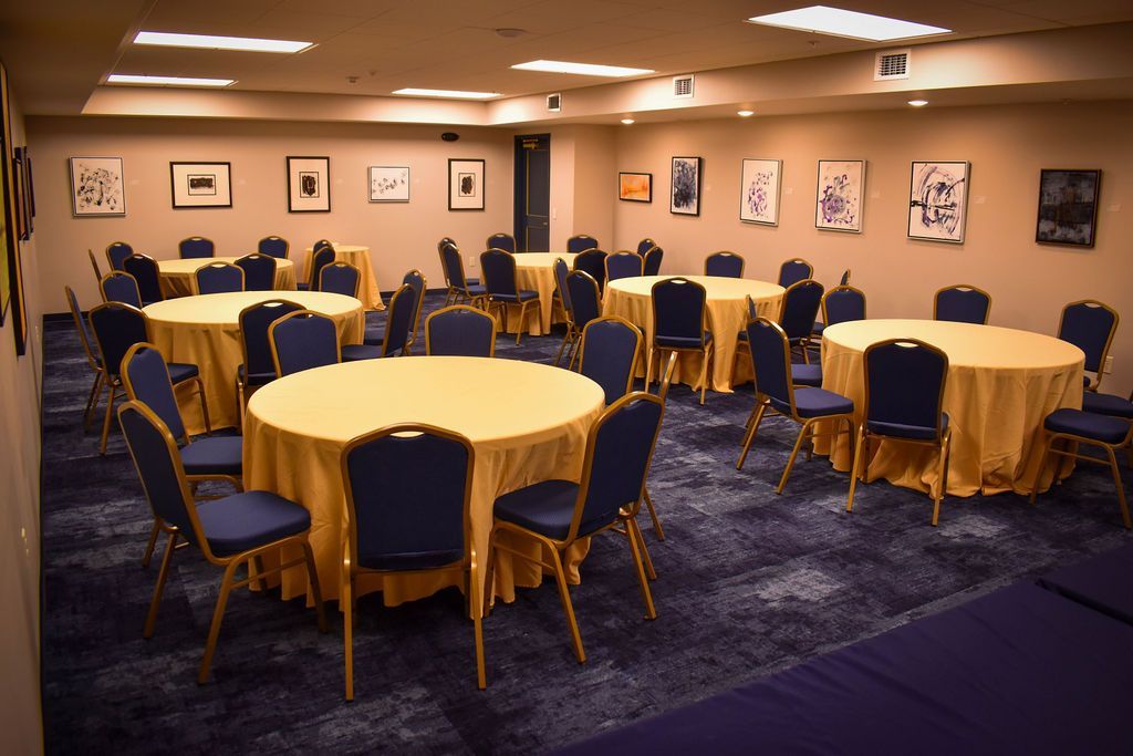 Empty event room with several round tables covered in yellow tablecloths, blue chairs, and art hung on tan walls.