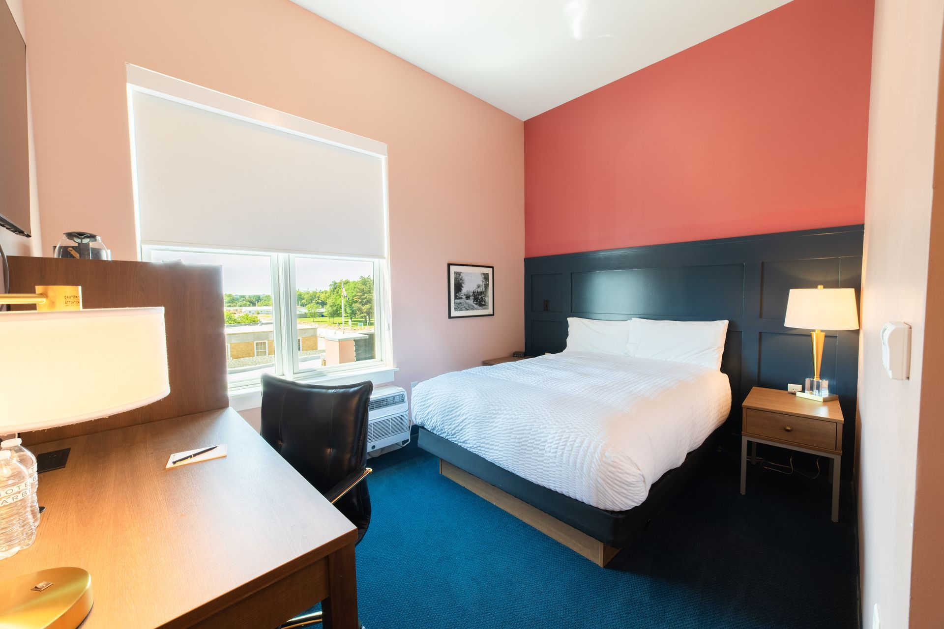 Hotel room with white bed, dark headboard, coral walls, blue carpet, and desk by a bright window
