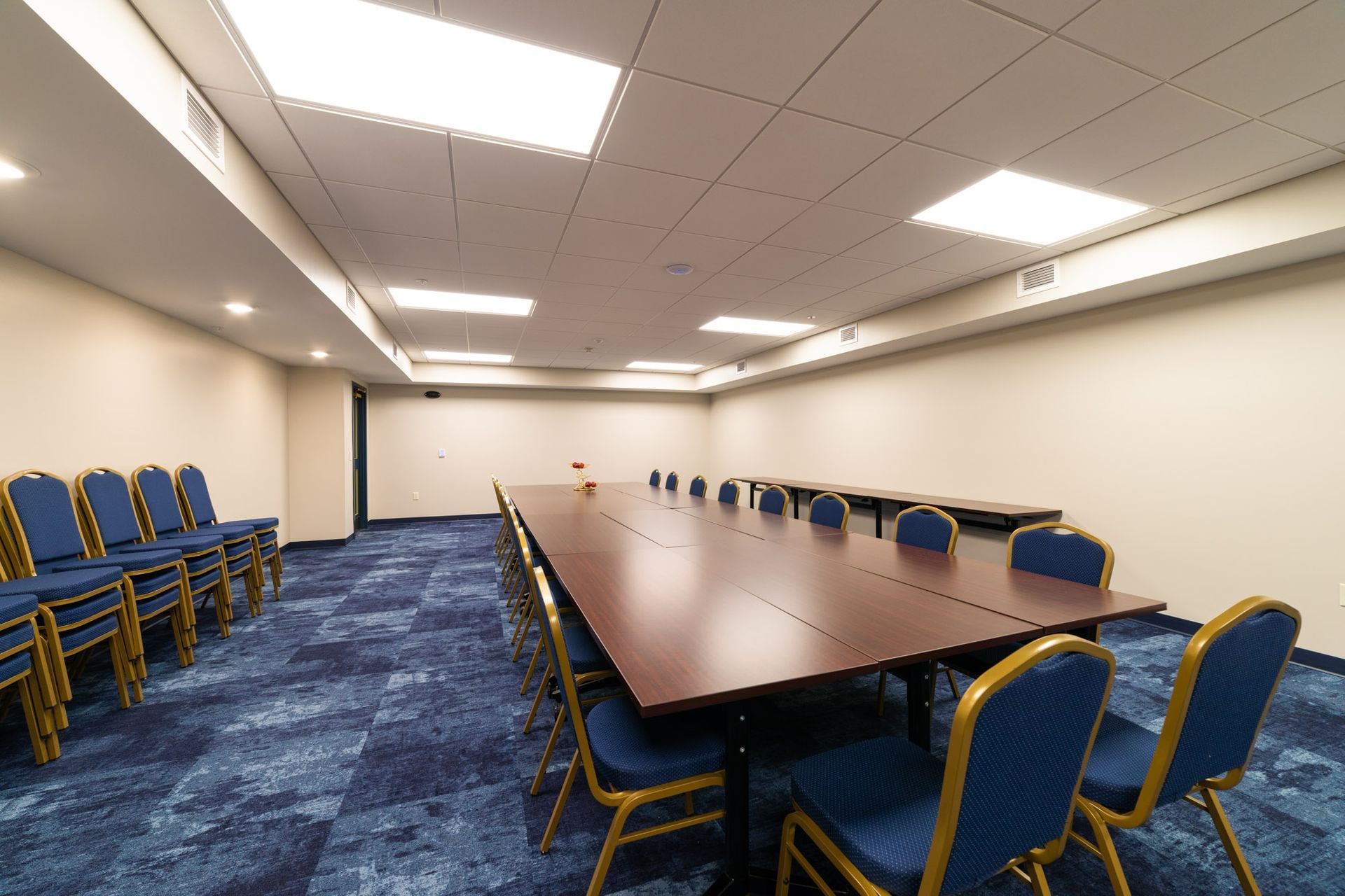 Conference room with long brown table, blue chairs, and blue carpet. Stacks of chairs on left.