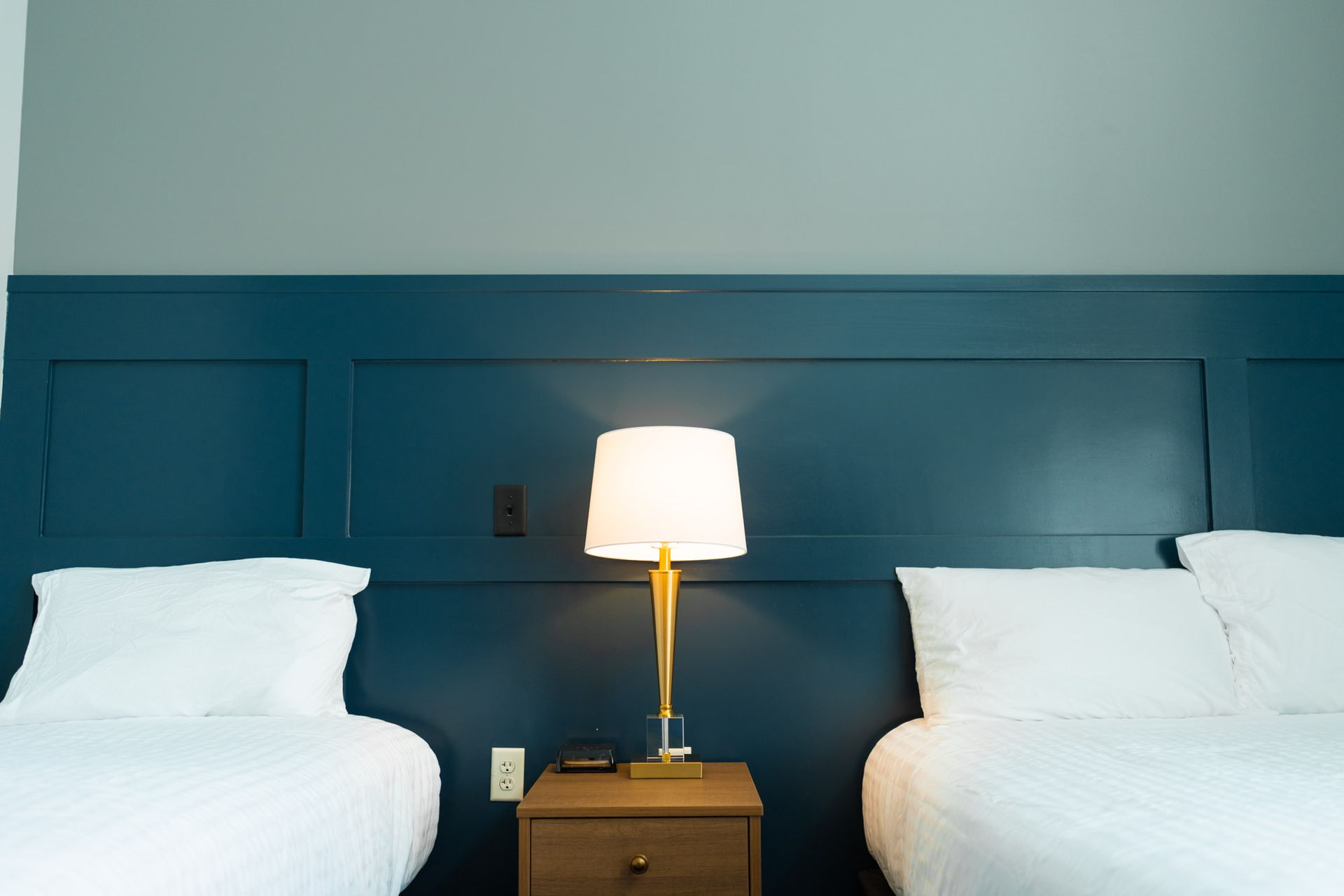 Two beds with white linens, a lamp on a nightstand, and blue paneling on the wall.