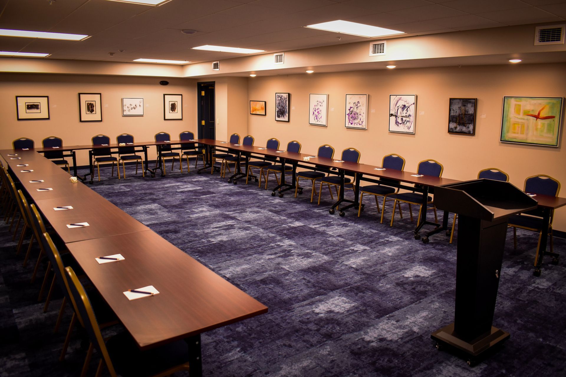 Conference room with tables arranged in a U-shape, blue carpet, art on beige walls, and a podium.