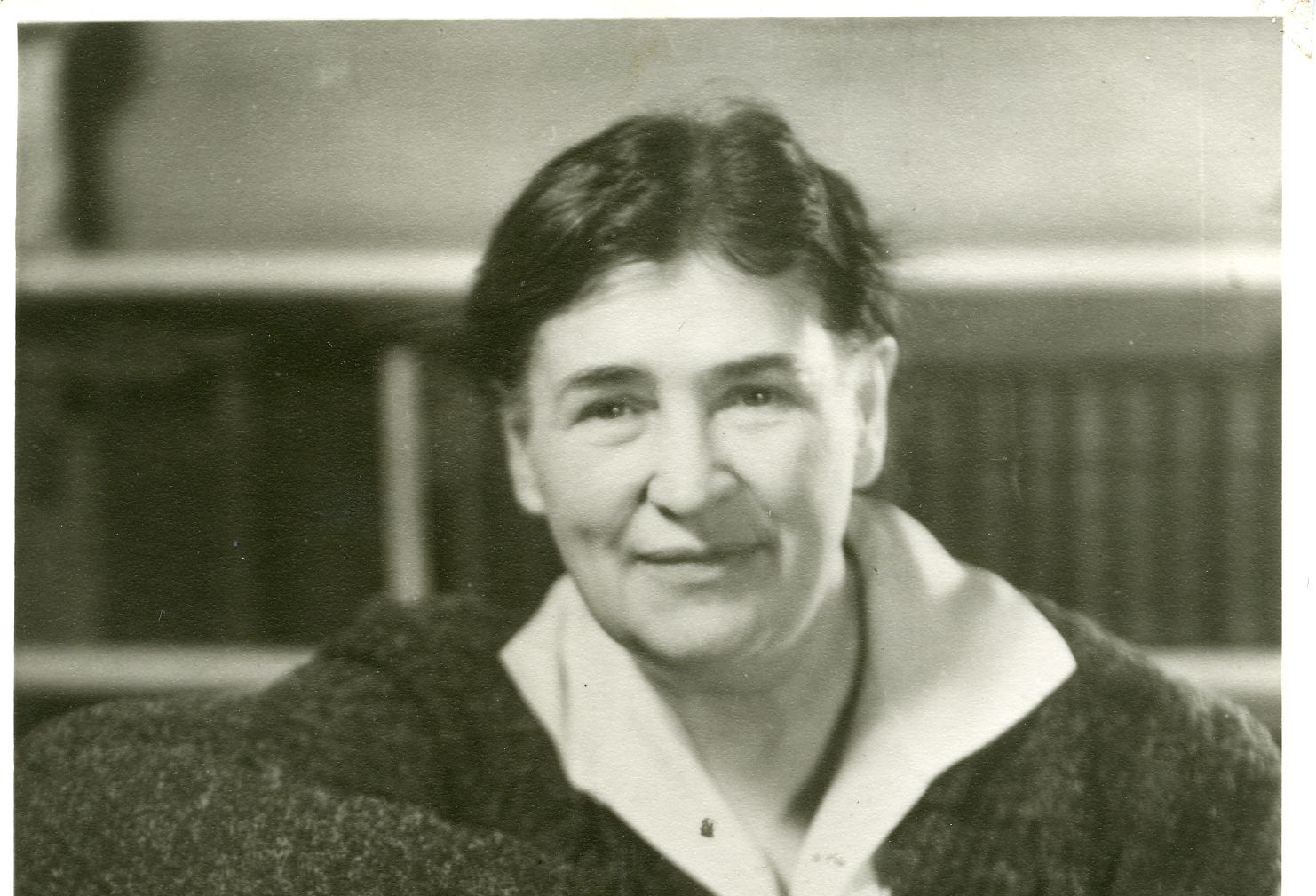 Woman with short dark hair, smiling, wearing a collared shirt and dark sweater. Bookshelf in the background.