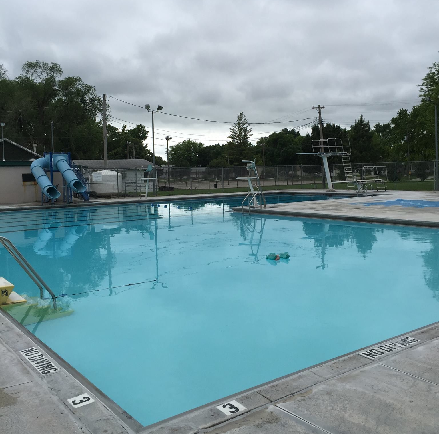 Outdoor swimming pool with clear blue water, diving board, slide, and cloudy sky overhead