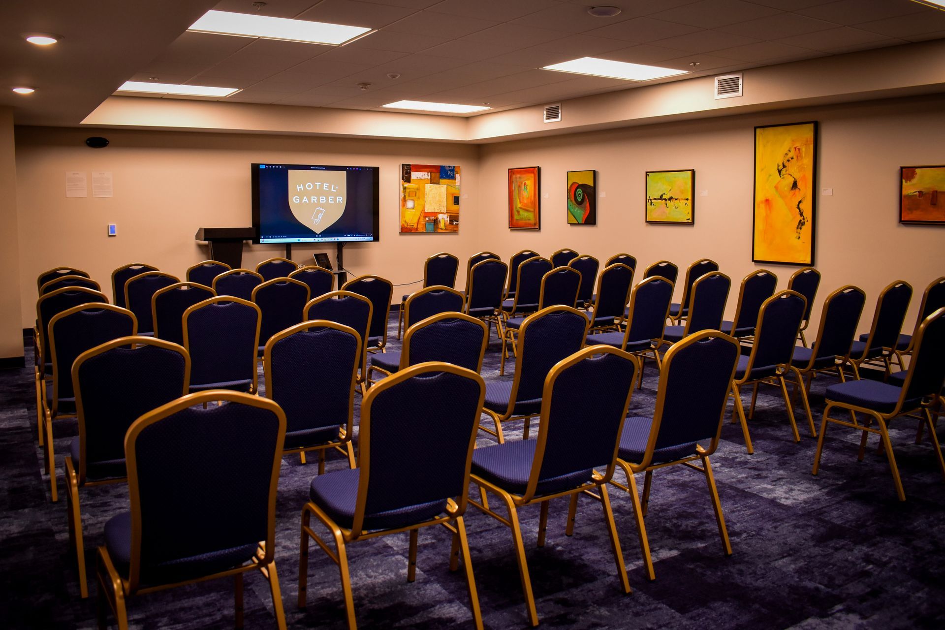 Empty conference room with blue chairs arranged for presentation, screen with logo, and art on walls.
