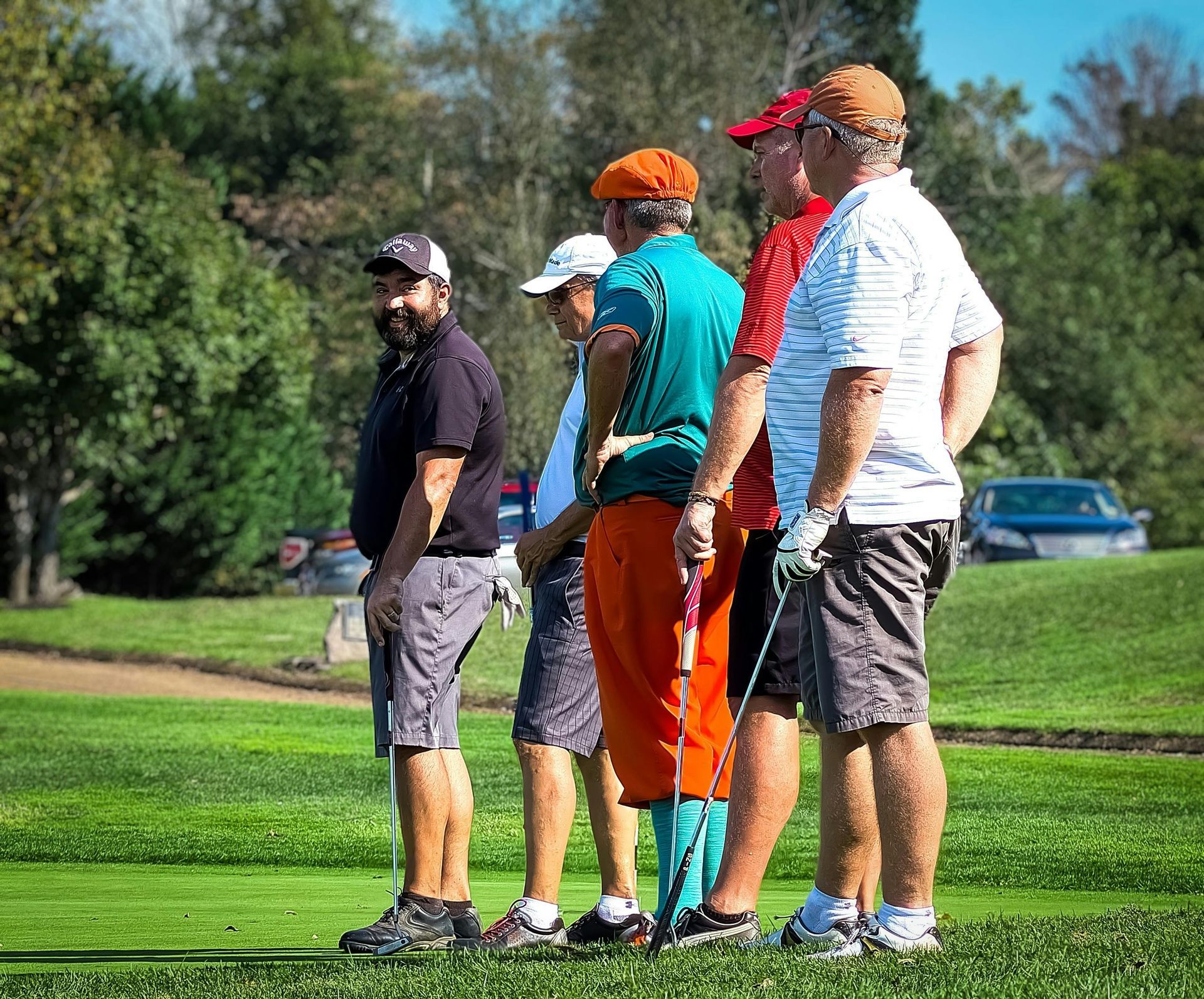 Five golfers stand on a lush green course, observing play. The person on the left looks toward the camera.
