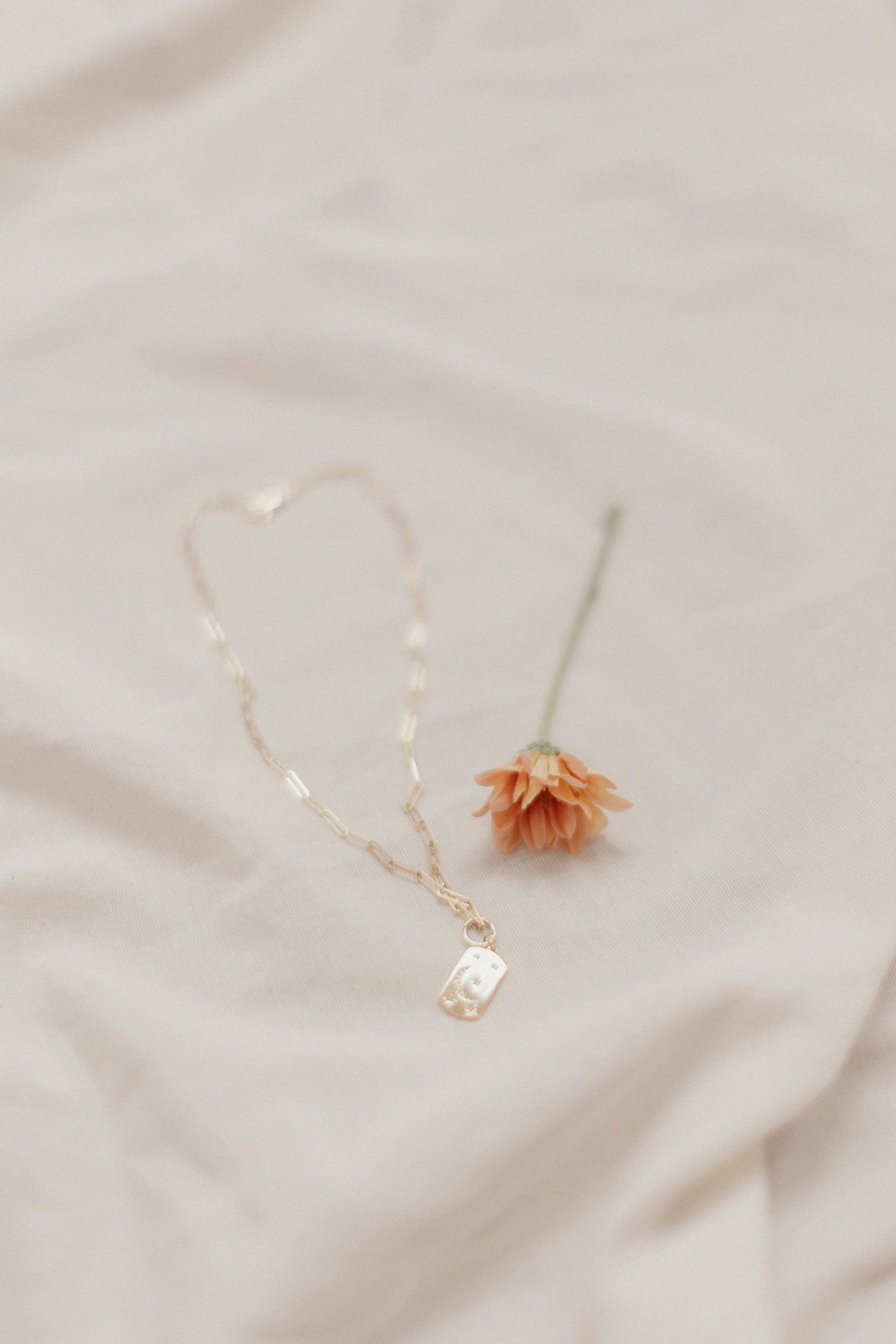 Gold necklace with small tag next to an orange dried flower on a cream-colored fabric.