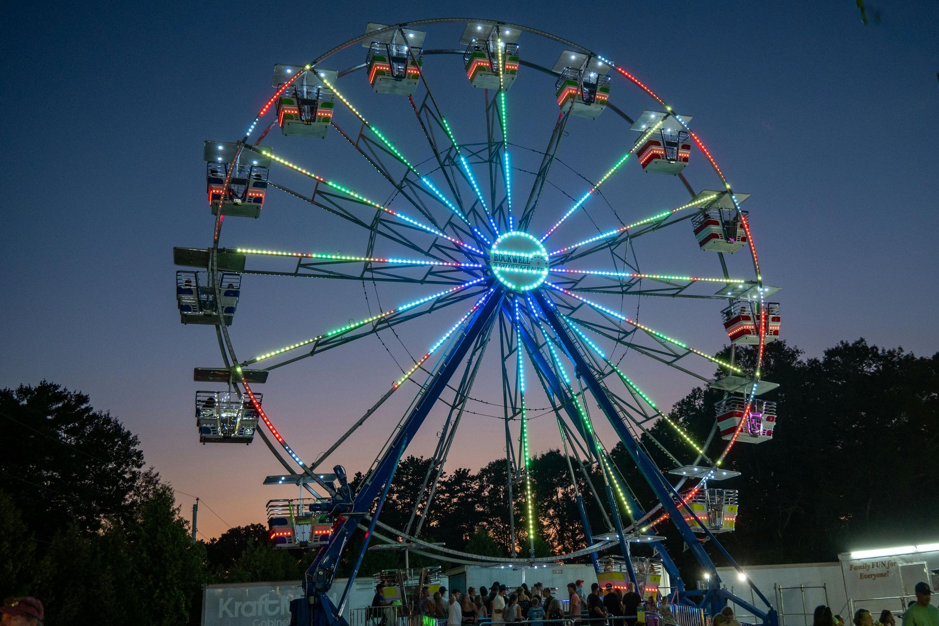 Ferris wheel lit with colorful lights at dusk; people gather below.