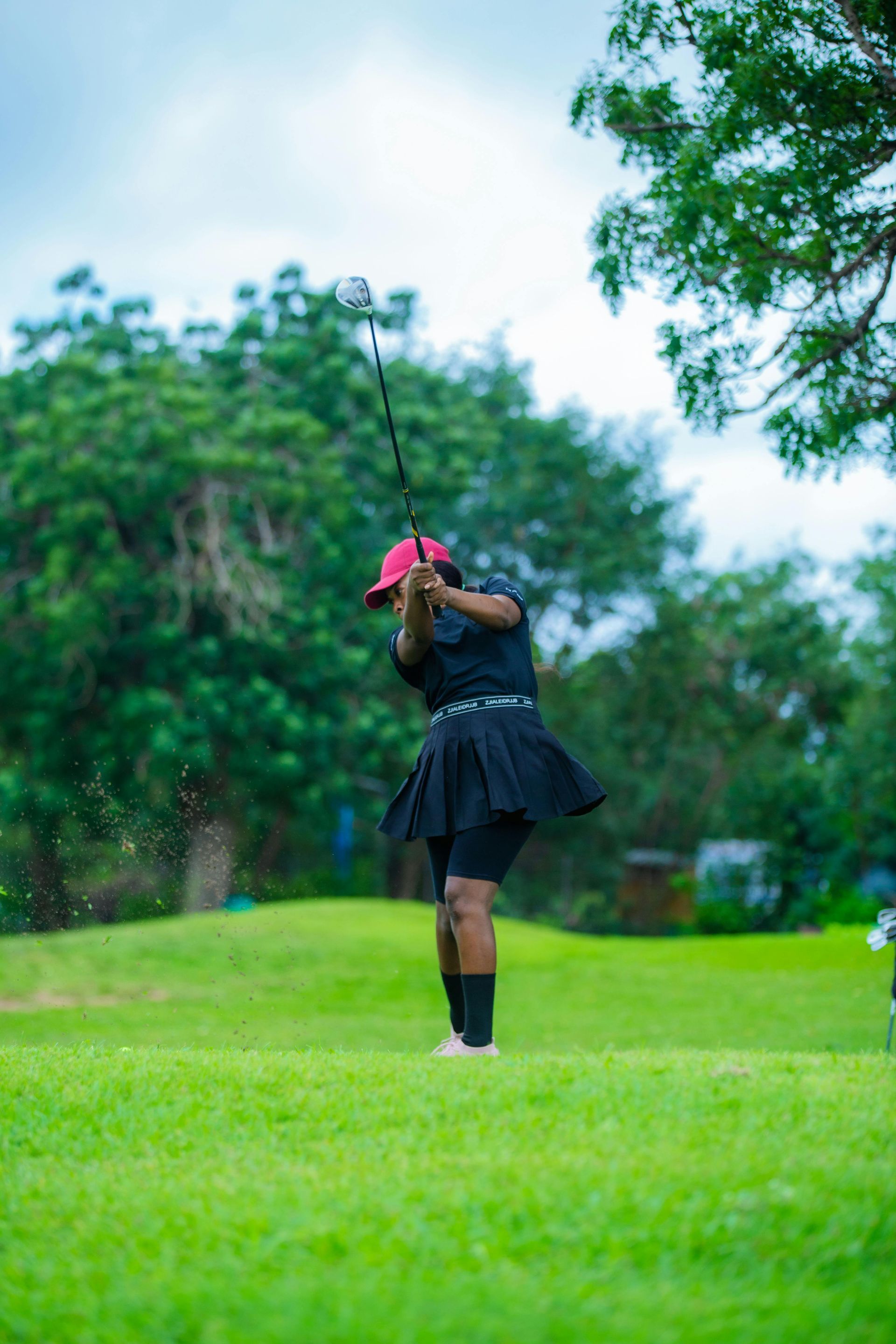 A person in a black outfit and red cap follows through on a golf swing on a bright green grass course with trees behind.