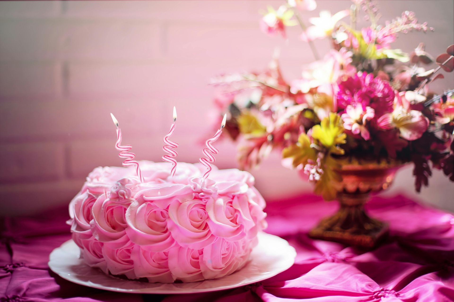 Pink frosted cake with three candles, next to a vase of flowers on a pink tablecloth.