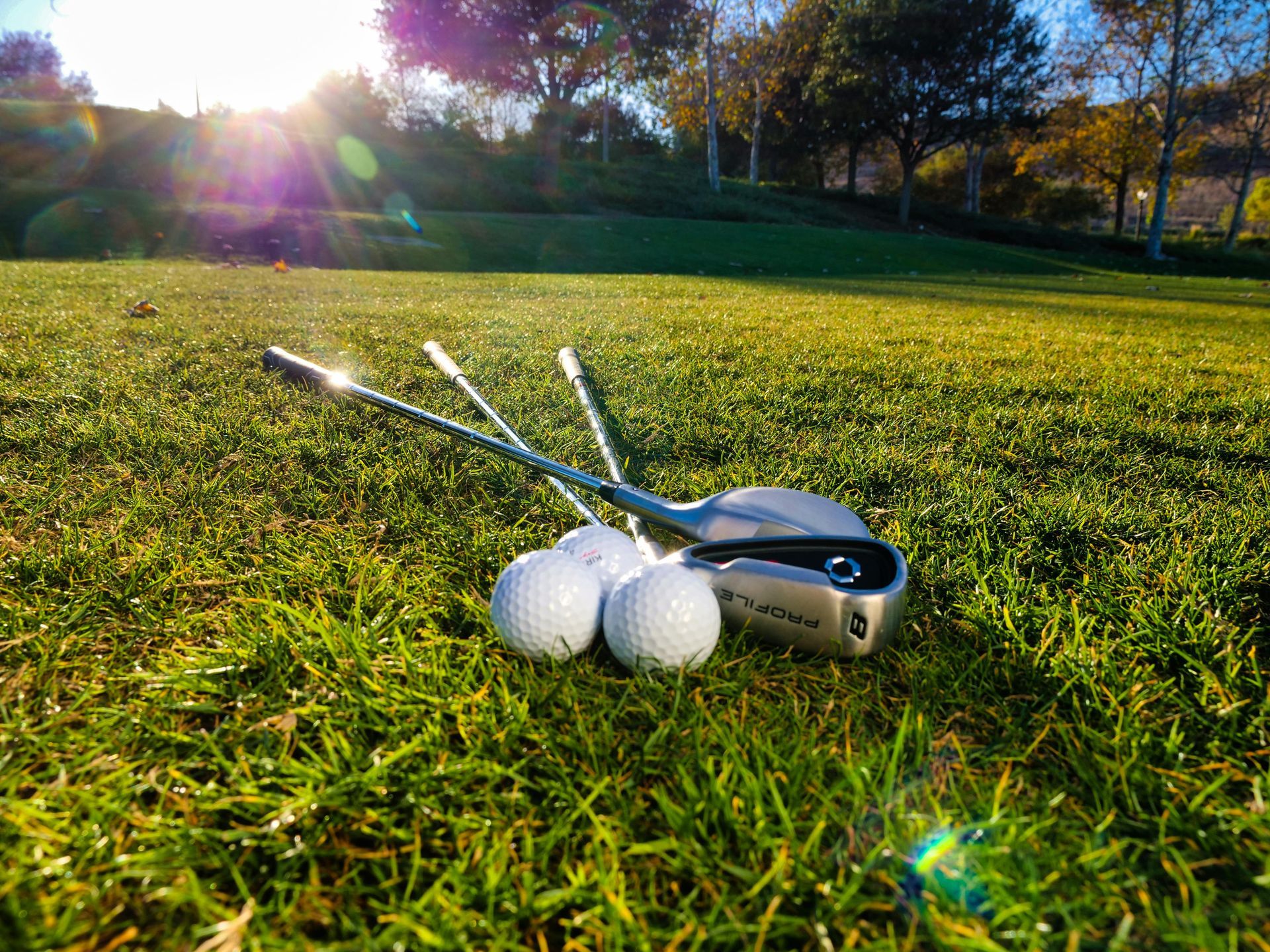 Three white golf balls and two golf irons rest on a grassy putting green at sunset.