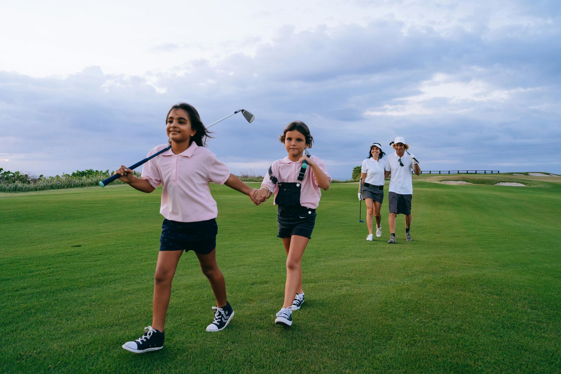 Two children walk hand-in-hand across a sunny golf course, with two adults walking behind them carrying golf gear.