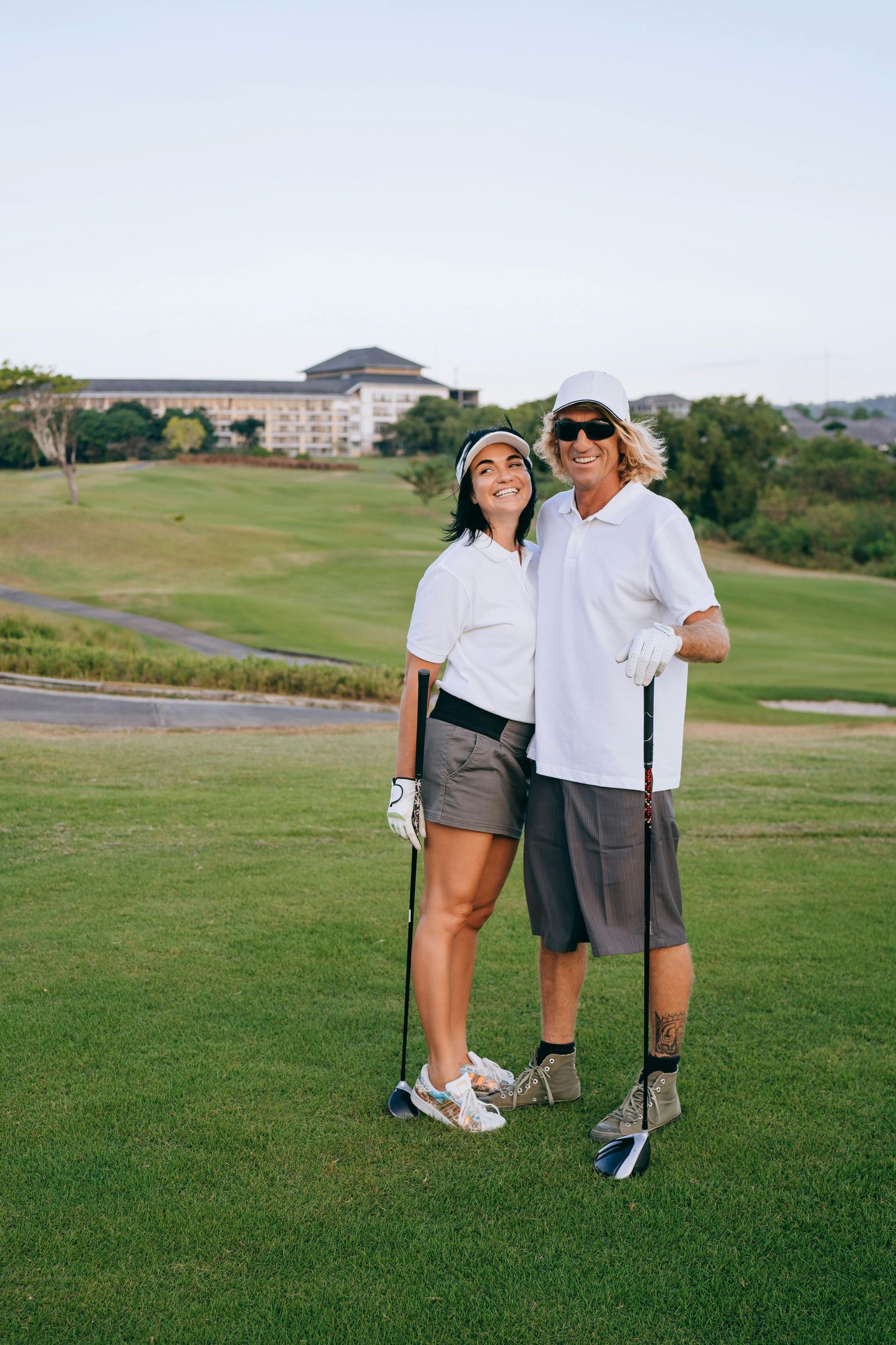 A couple dressed in white shirts and shorts stands on a green golf course, holding clubs and looking up at the sky.