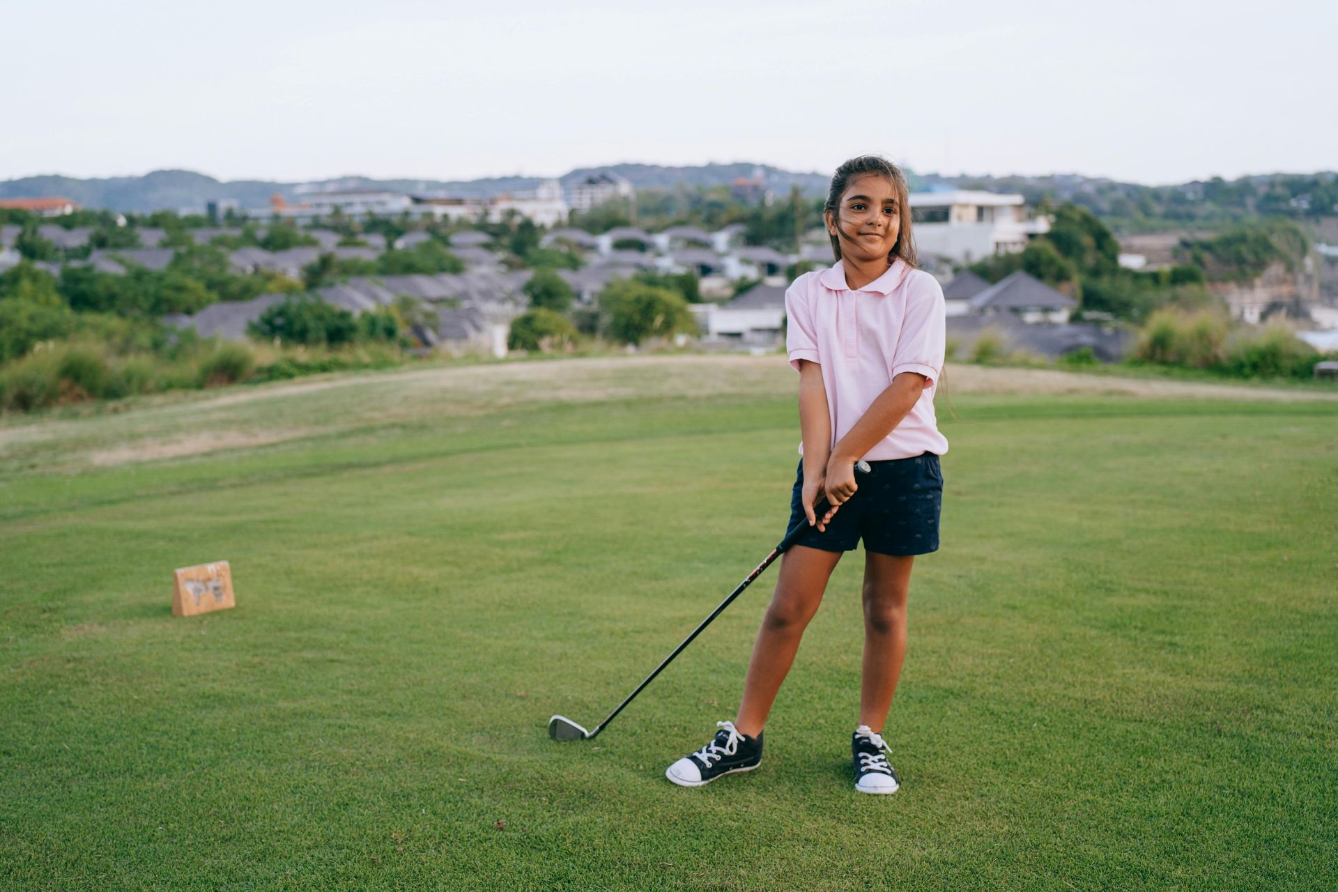 A young person in a pink polo and dark shorts holding a golf club on a grassy course with a residential view in the back.