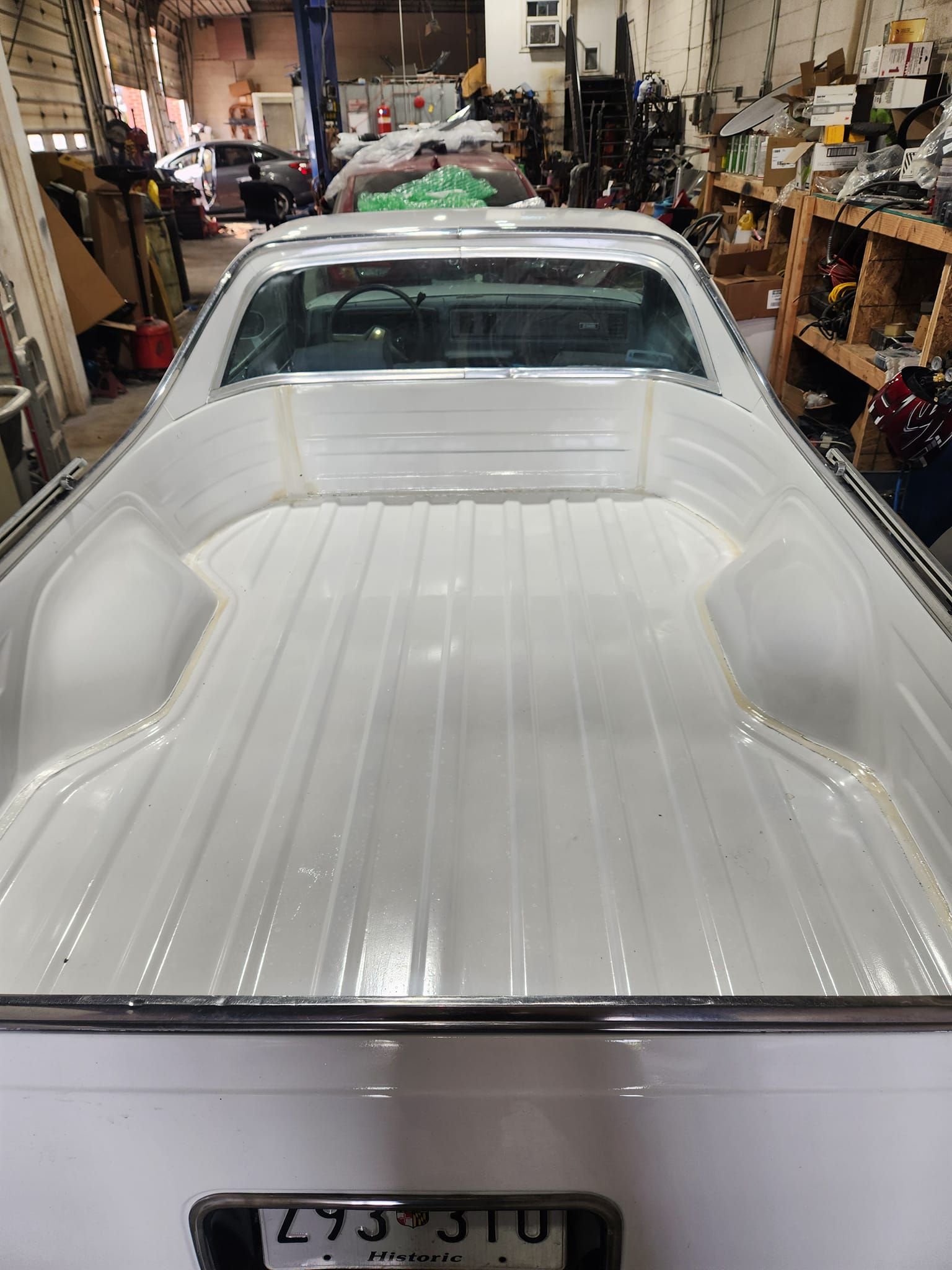 White car being restored in a cluttered garage, viewed from the rear with the bed or trunk exposed.