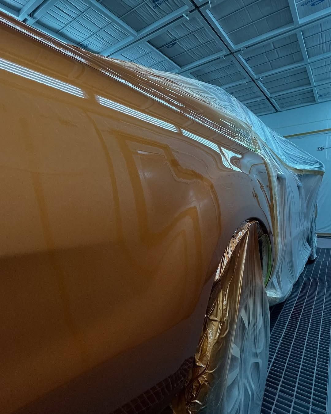Orange car body panel in a spray booth, with masking and protective sheeting around the wheel area.