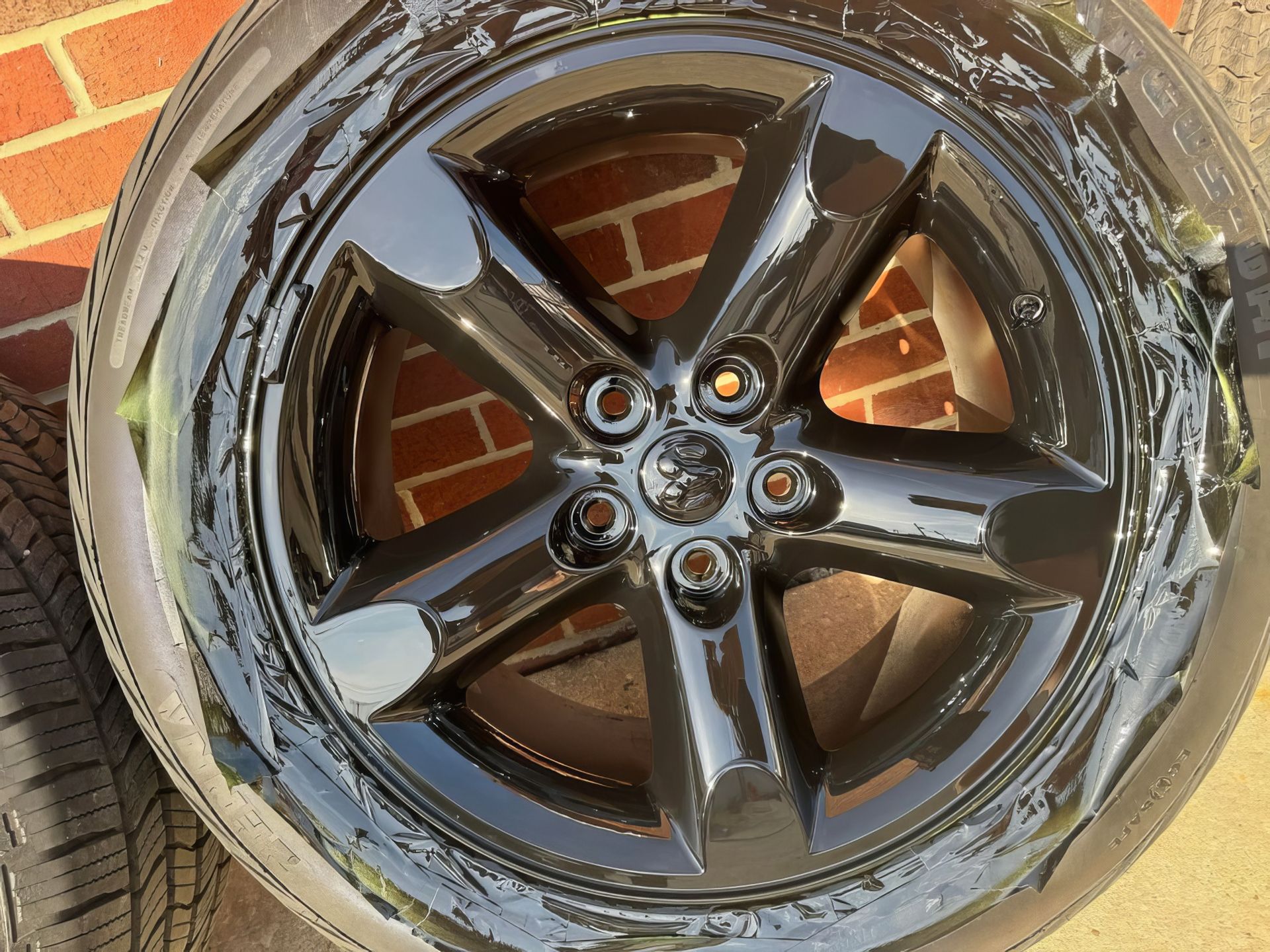 Glossy black five-spoke car wheel with wet tire, close-up against a brick wall and pavement