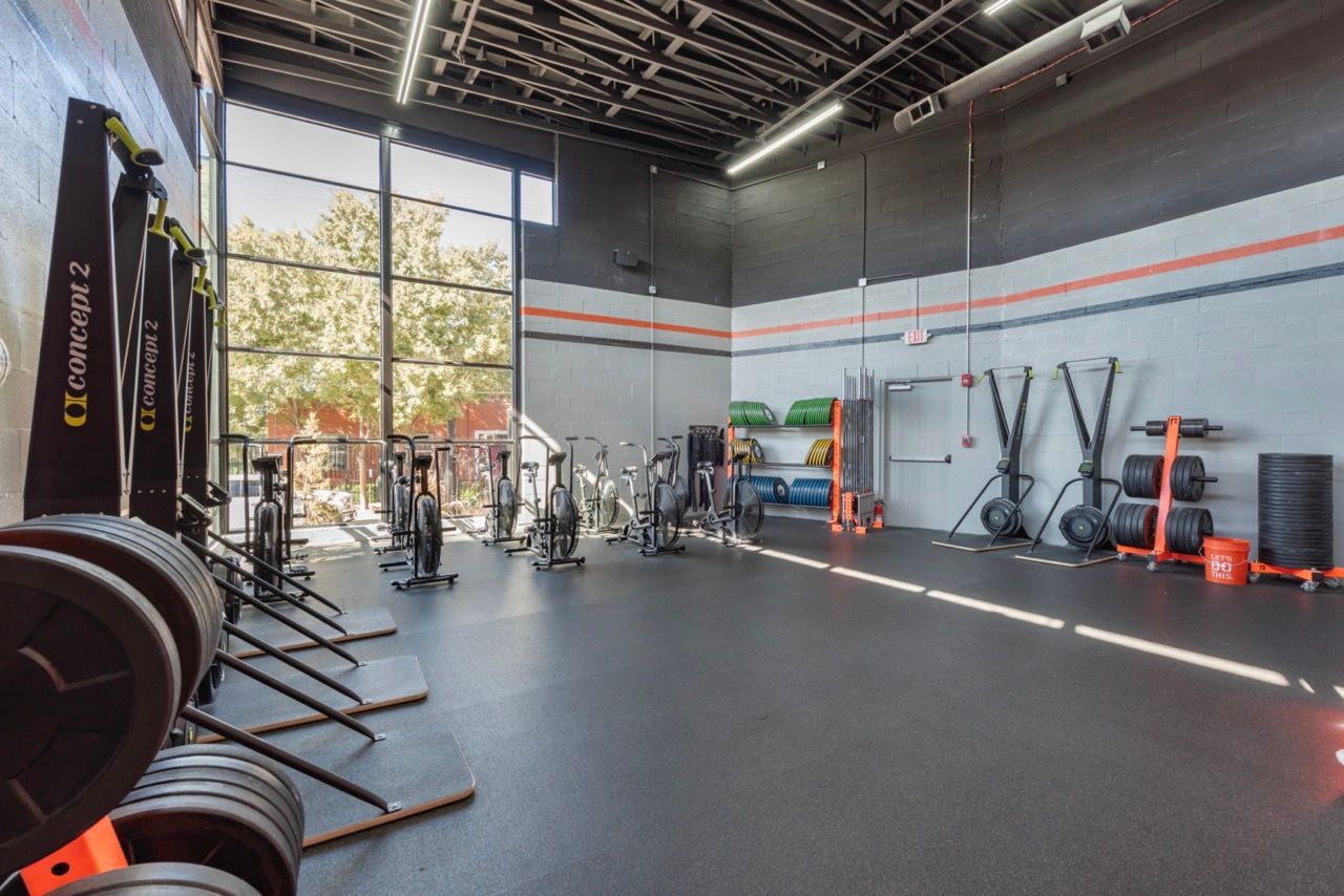 Gym interior with exercise equipment, including weights, bikes, and rowers. Large window, dark gray walls.