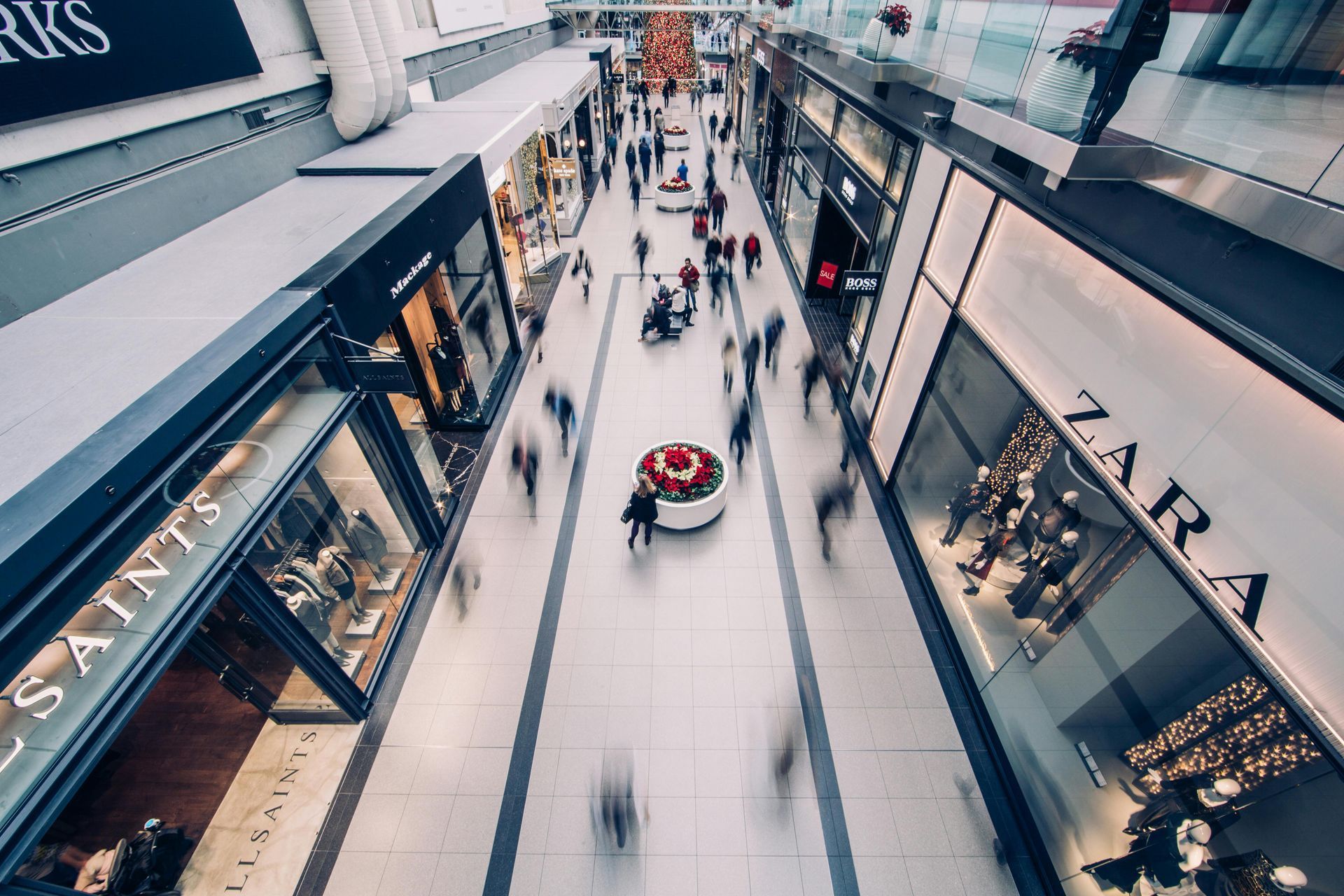 Mall interior, many blurred people walking. Stores line both sides, some with displays; Zara visible.