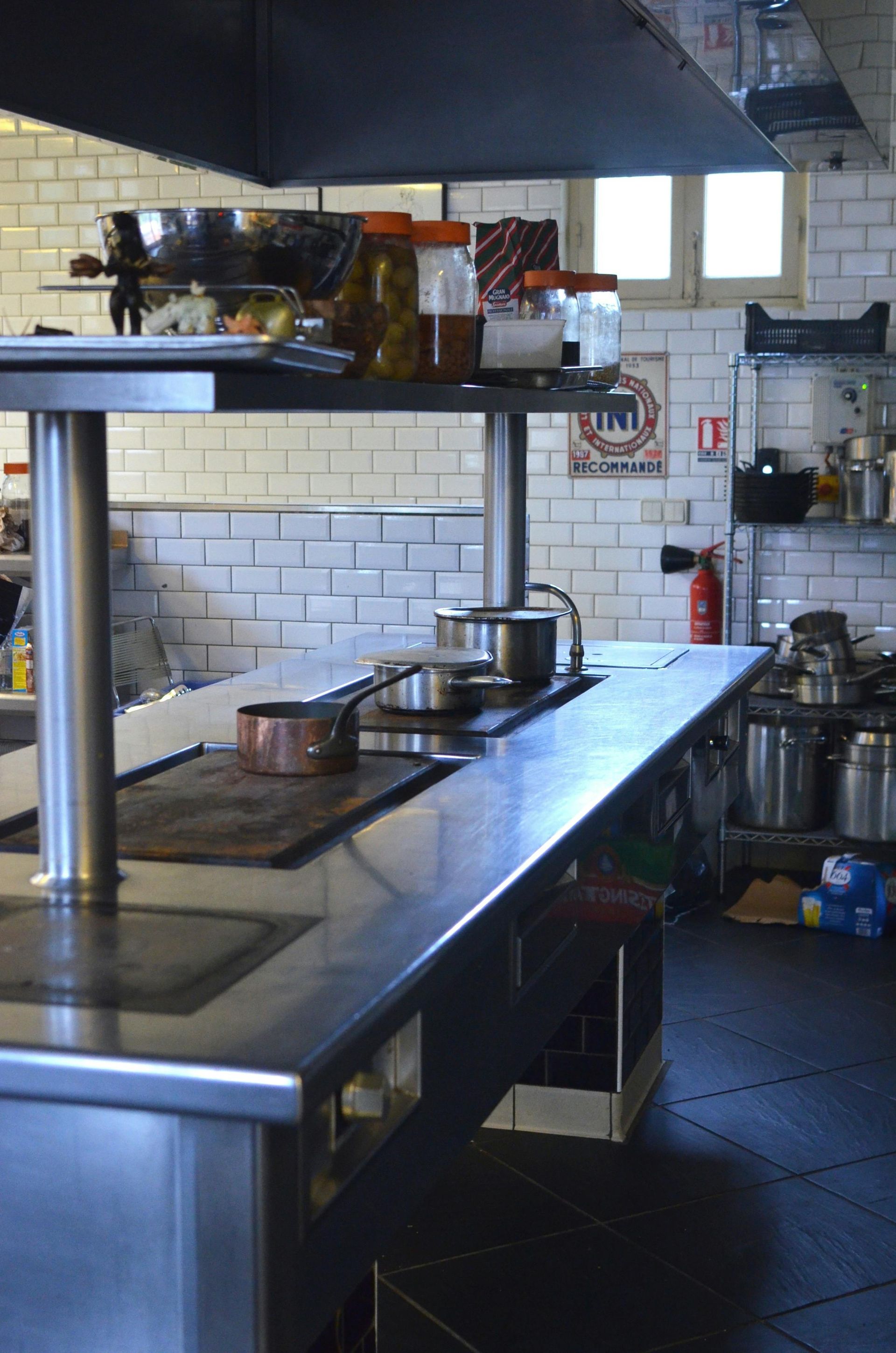 A commercial kitchen with stainless steel countertops, stovetop, and cookware. White tile walls.