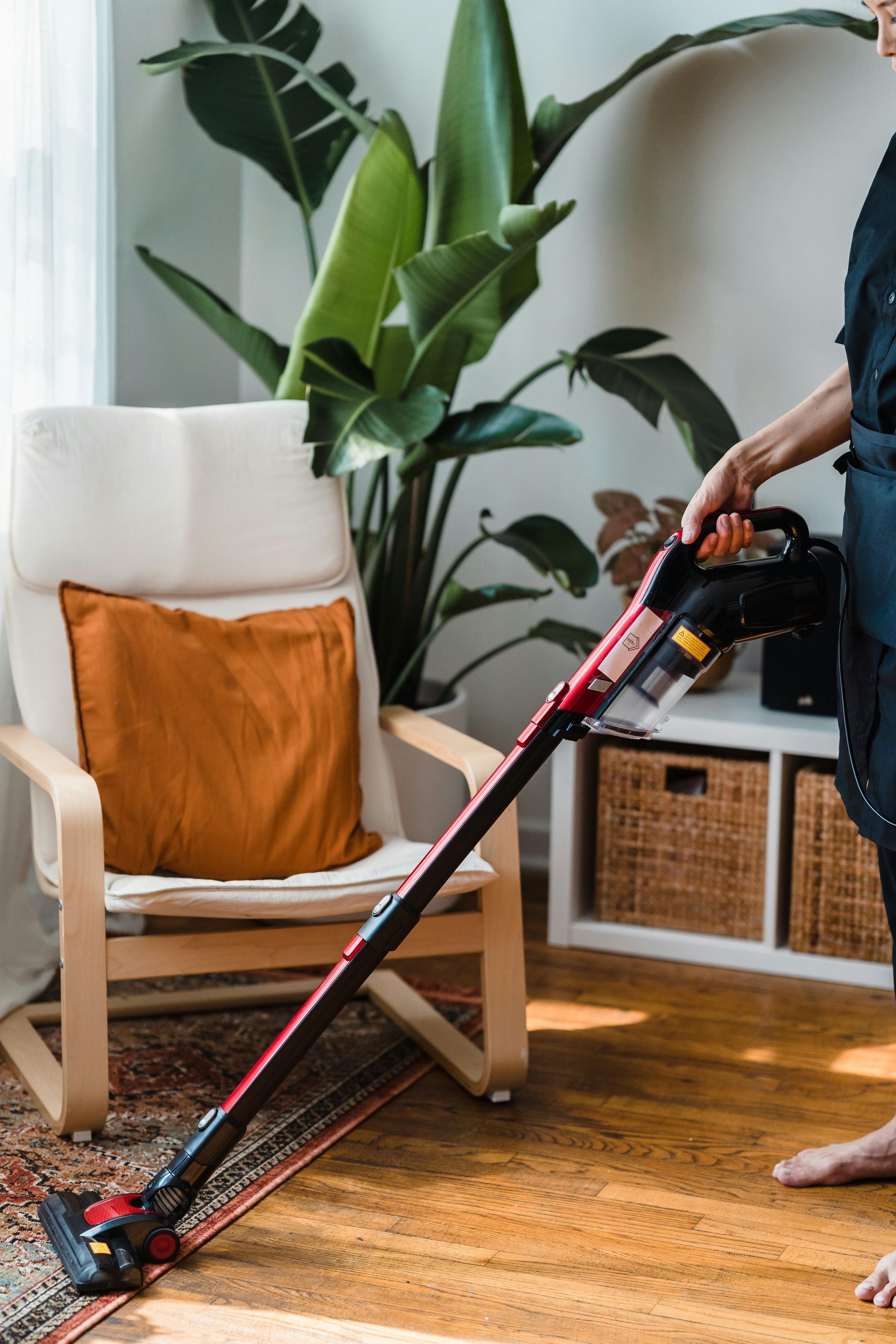 Person vacuuming wooden floor, near a chair with an orange pillow and a houseplant.