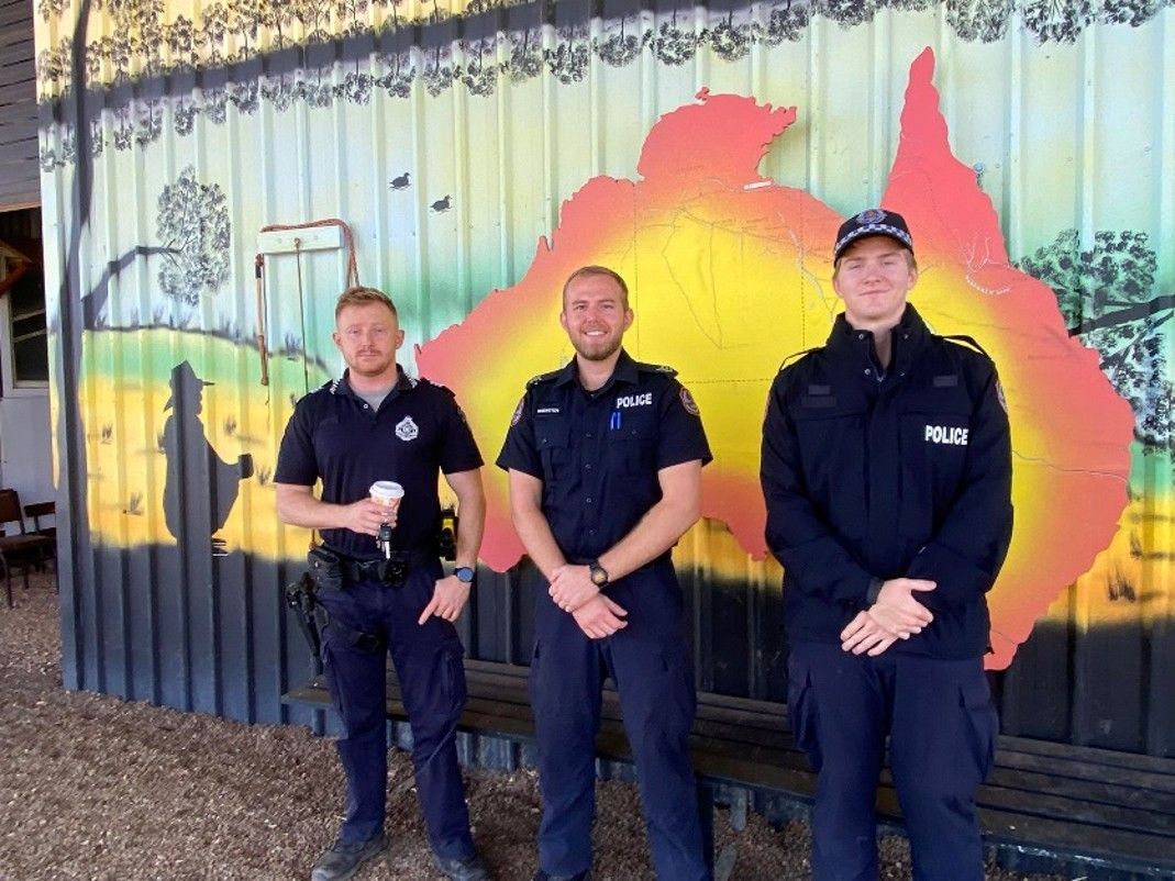 Three uniformed officers standing in front of a colorful mural with a sunset and city skyline.