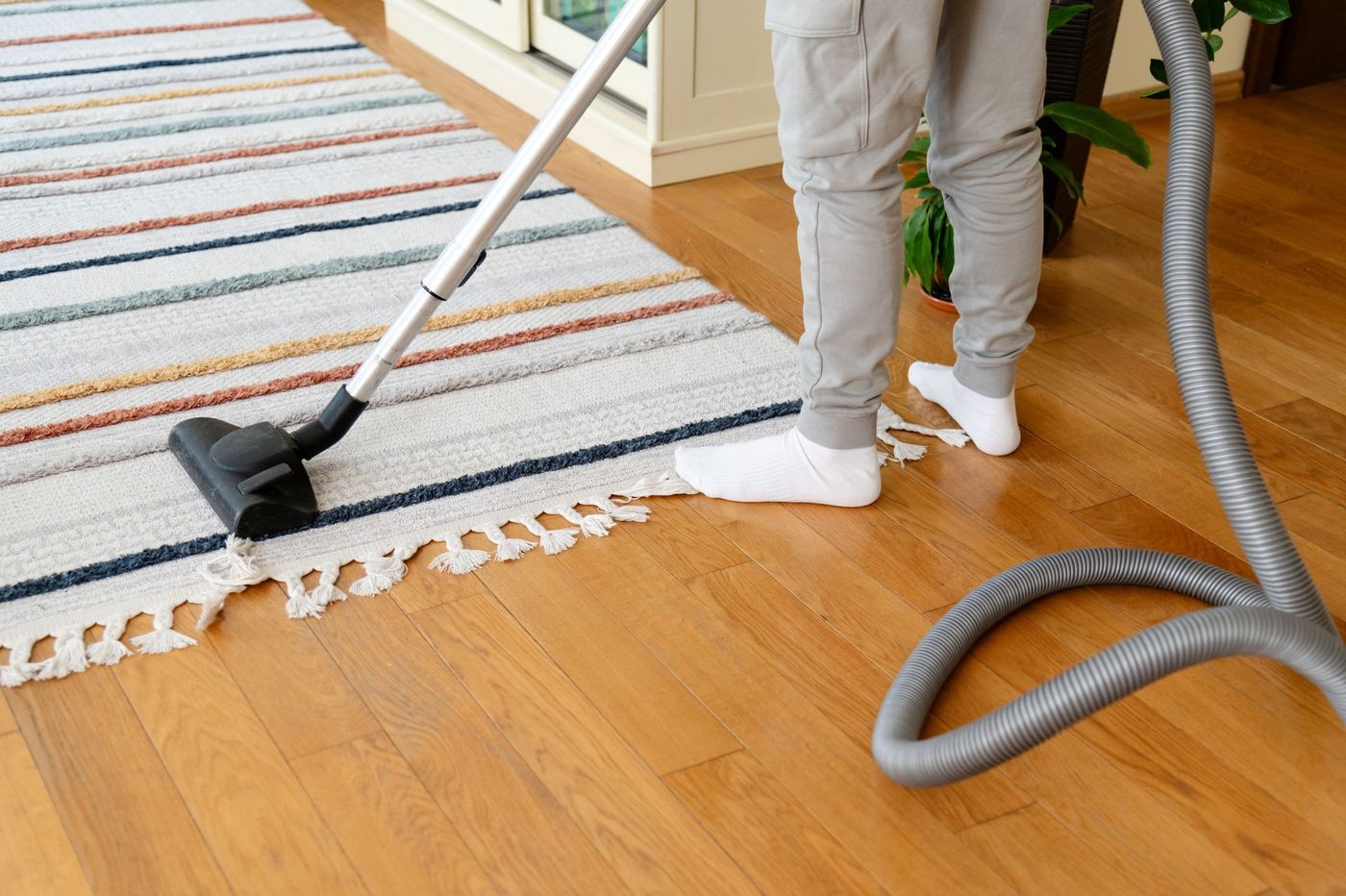 Person vacuuming a striped rug beside a hardwood floor, with a vacuum hose coiled nearby.