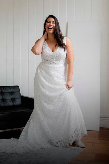 Woman in a white lace wedding dress laughs. Leaning on a black couch in a white room.