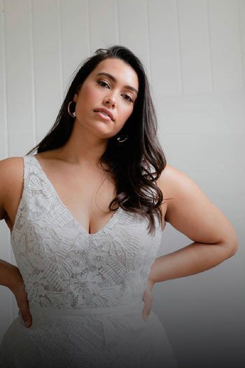 Woman in a white lace dress poses with hands on hips, looking towards the camera.