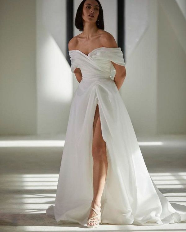 Woman in off-the-shoulder white wedding gown with high slit, standing in a bright, modern space.