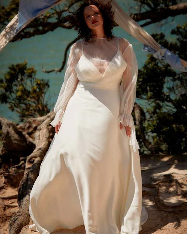 Woman in white wedding dress, sheer sleeves, standing outdoors, coastal setting.