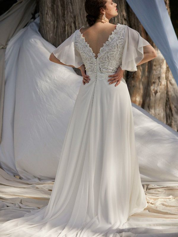 Woman in white wedding dress with lace bodice, butterfly sleeves, and flowing skirt, standing outdoors.