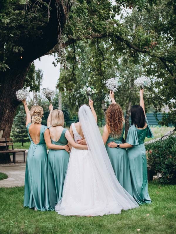 Bride and bridesmaids in teal dresses with bouquets, arms raised, under a tree.