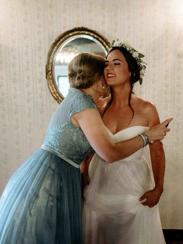Woman in blue dress hugs bride in white dress, near a mirror.