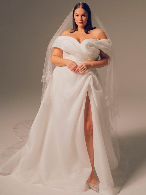 Woman in white off-shoulder wedding dress with high slit, wearing a veil, standing in a studio.