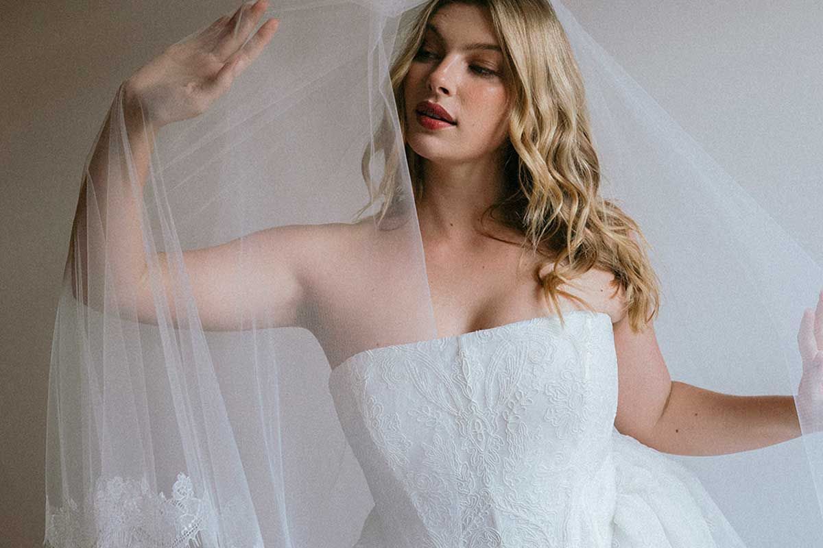 Bride in strapless white wedding dress, lifting veil, with blond wavy hair.