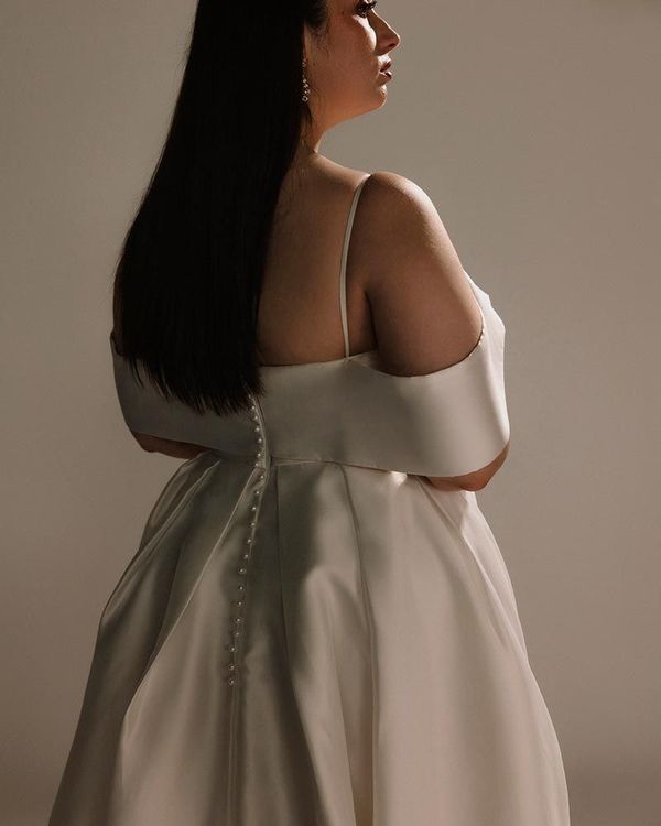 Woman in white off-the-shoulder wedding gown, standing, with long dark hair. Backlit against a white background.
