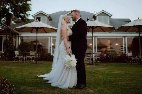 Bride and groom facing each other on grass, near a venue with umbrellas, looking at each other.