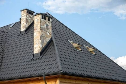 Dark gray roof with two stone chimneys and dormer windows against a blue sky.