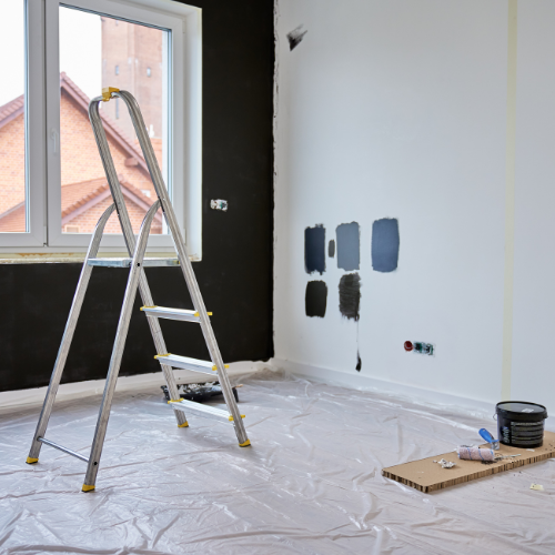 Room with ladder, half painted black and white, paint samples on wall, plastic floor covering.