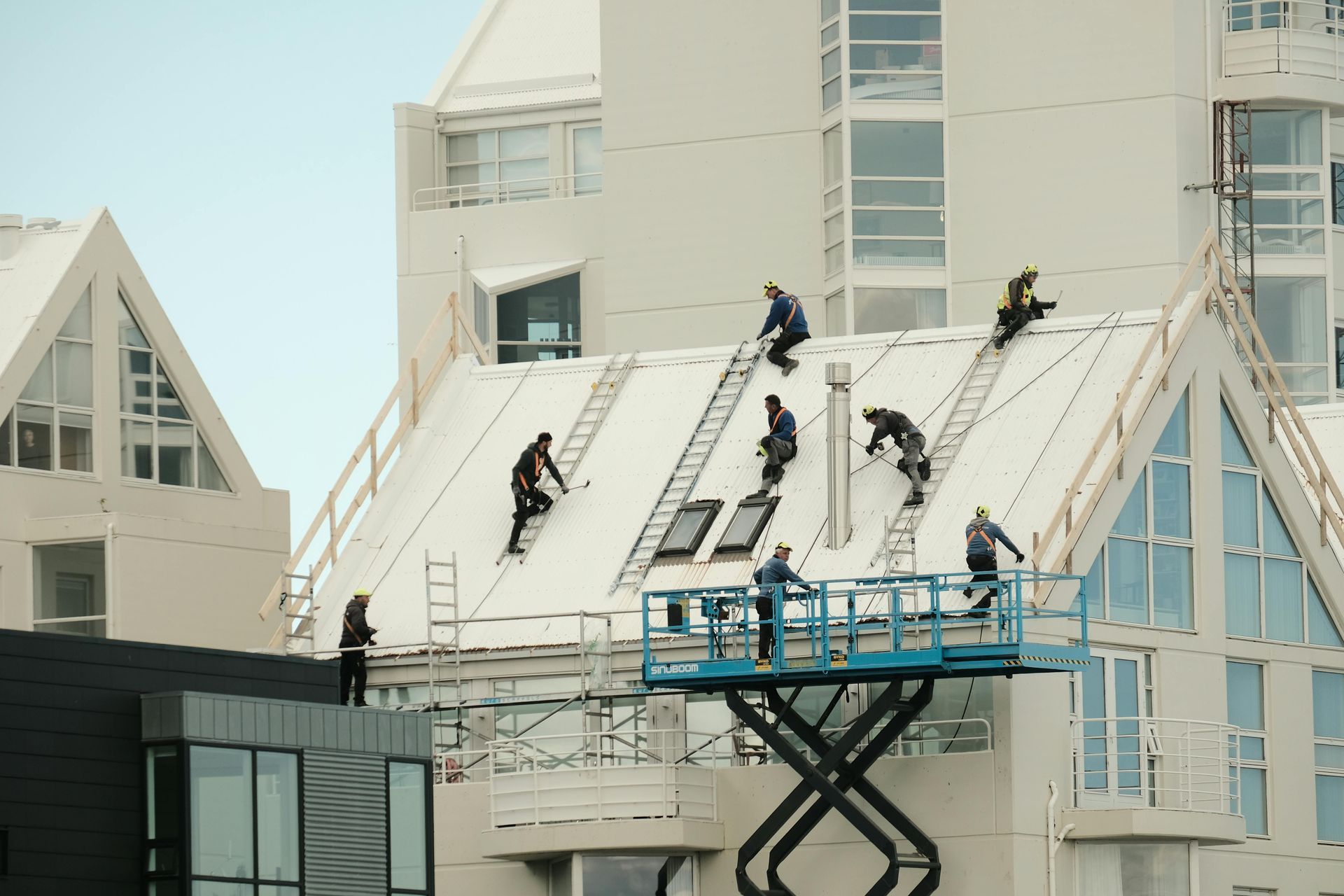Workers on a white building roof with ladders; several on a lift below. Cloudy sky.