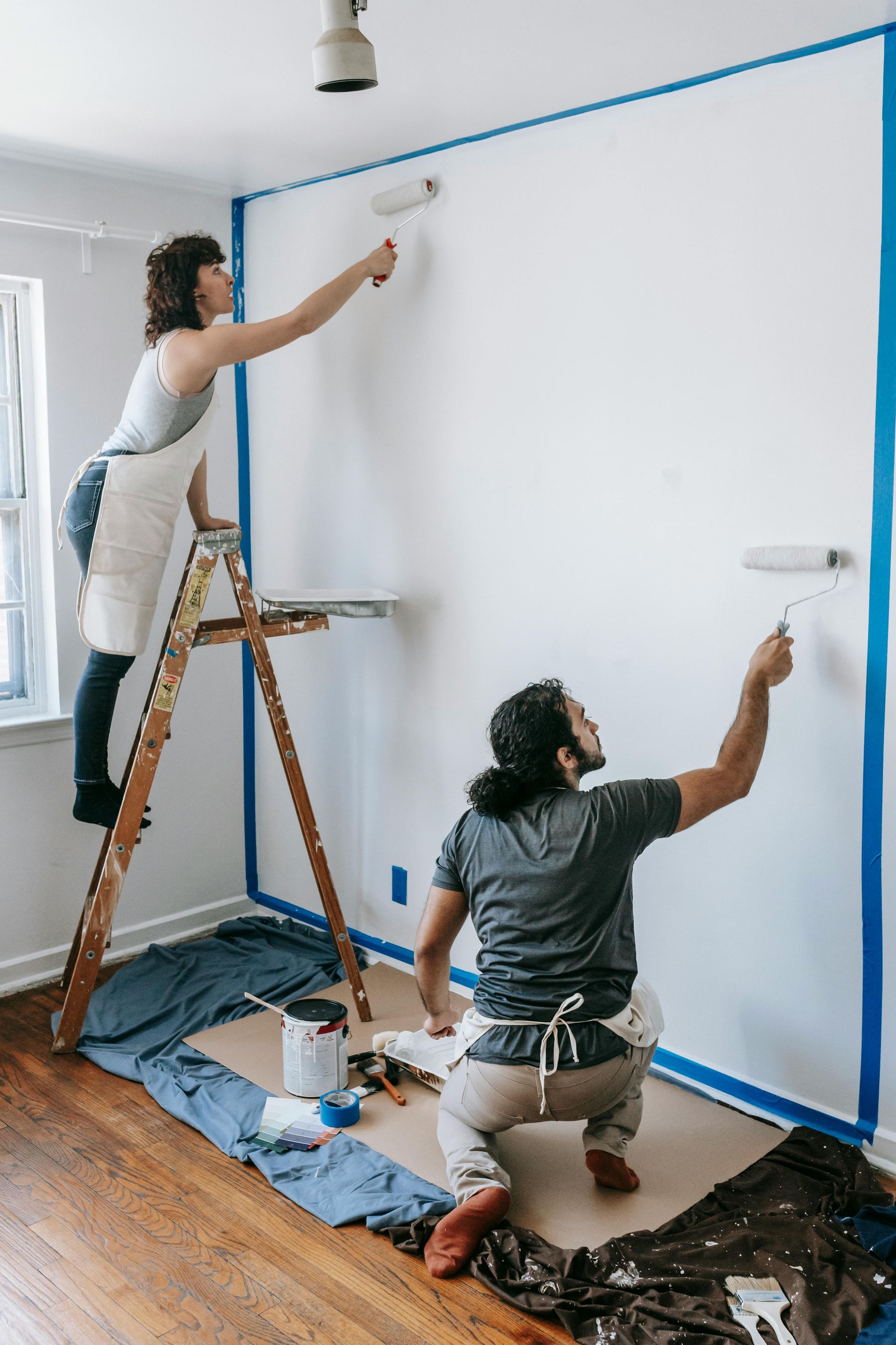 Two people painting a wall: one on a ladder, the other kneeling. Blue painter's tape outlines the wall. 