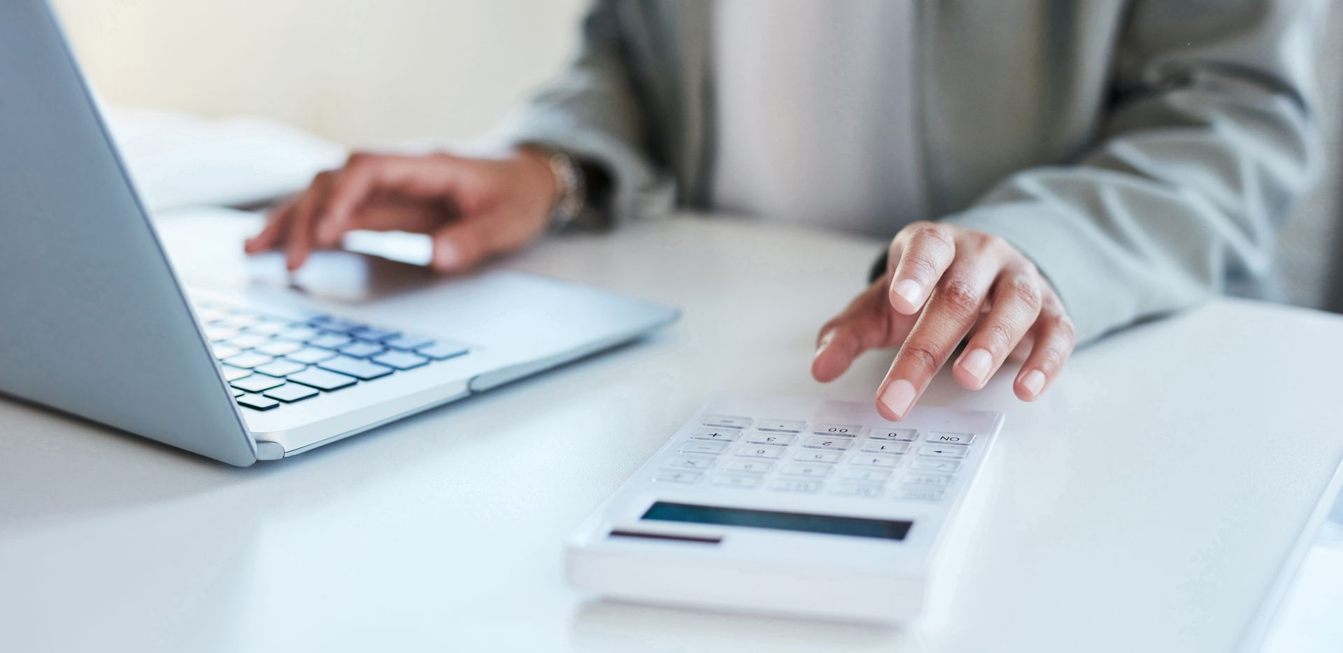 Person using a laptop and calculator on a white desk, hands visible.