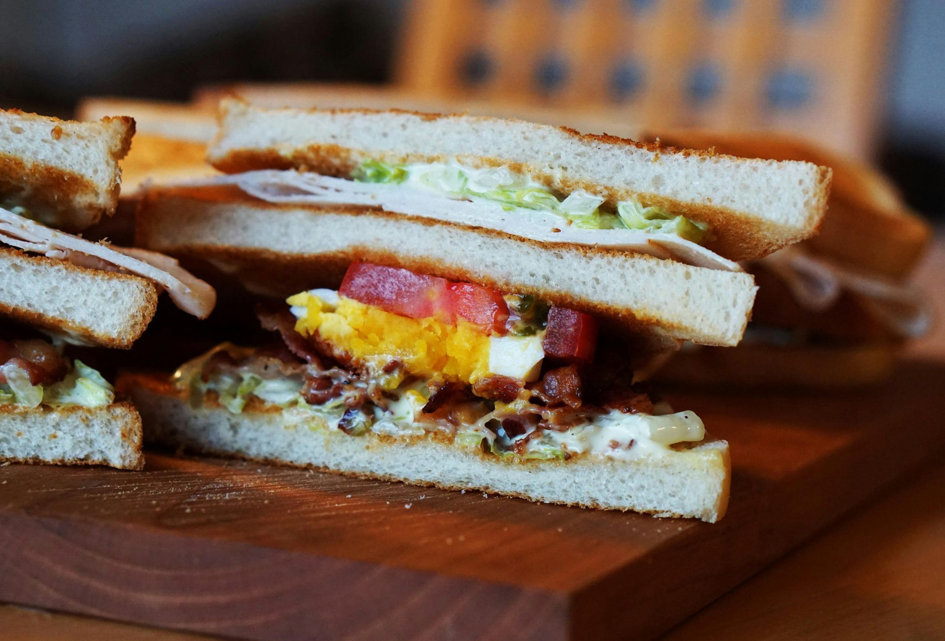 Club sandwich on a wooden board, layers of ingredients visible: bacon, egg, tomato, lettuce, turkey, and toasted bread.
