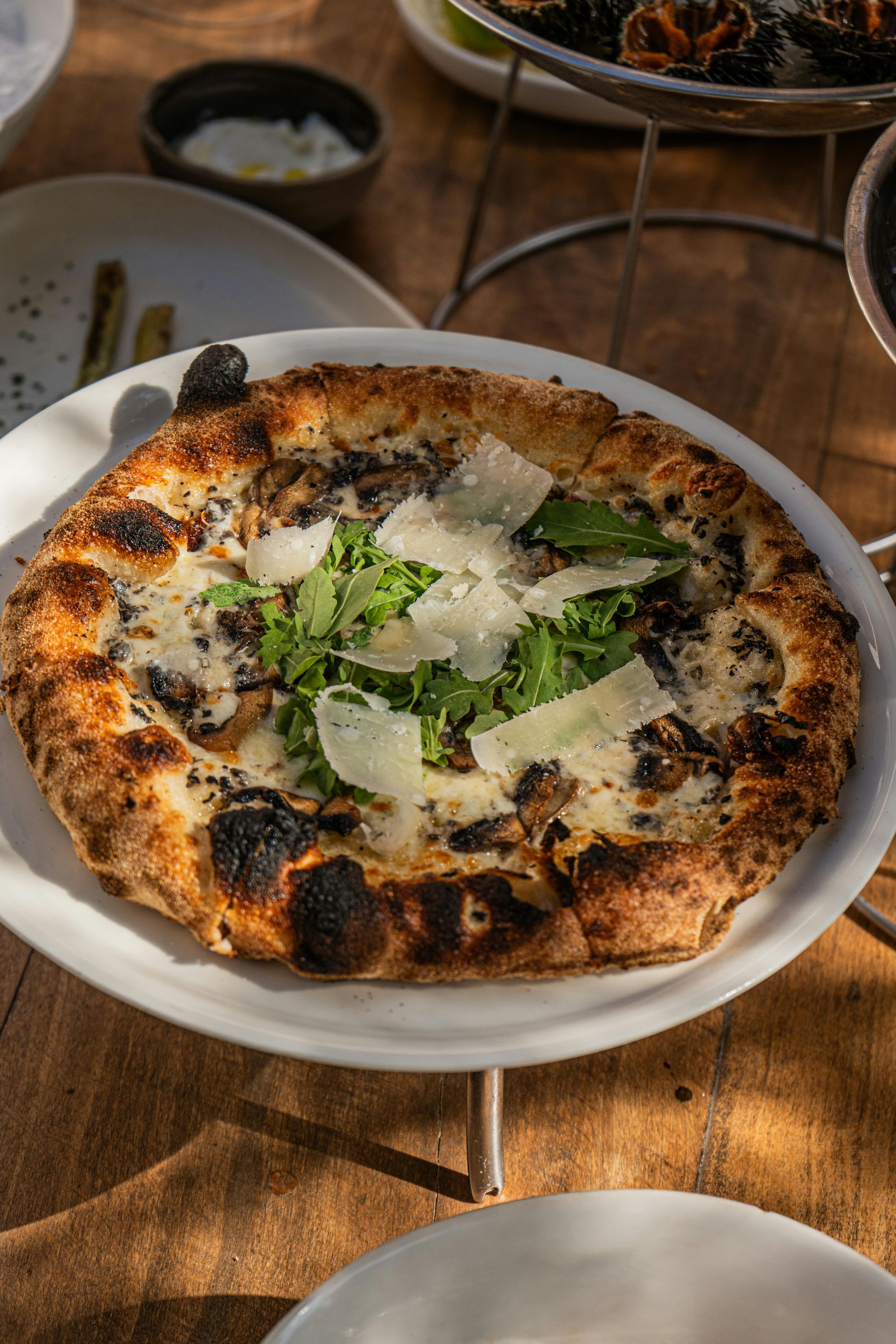 Pizza on a white plate, topped with greens and shaved cheese, set on a wooden table.